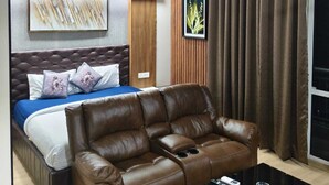 A modern living room with a brown leather sofa, a bed with a blue bedspread, a ceiling fan, and a large window with curtains. - Homeiihood Stay Lucknow (Lucknow)