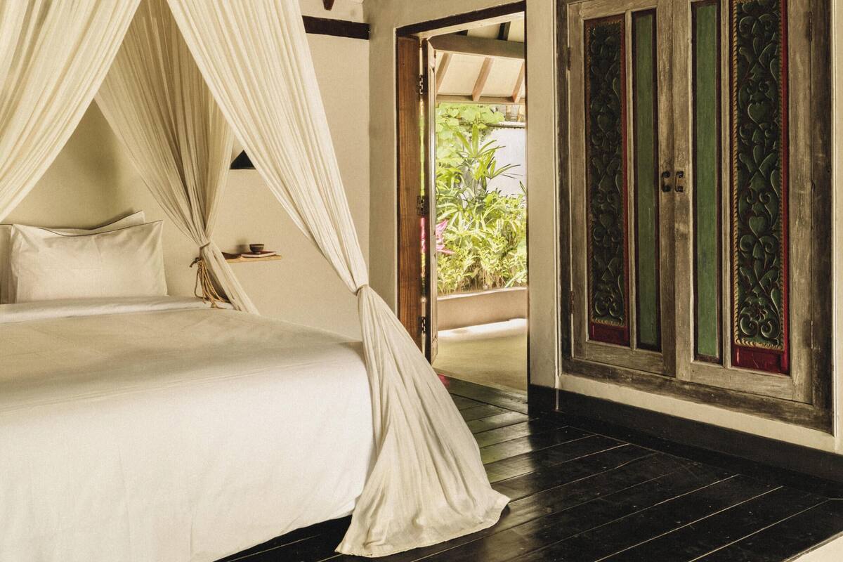 A bedroom with a canopy bed, a wooden wardrobe, and a view of greenery outside.