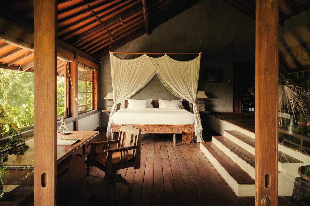 A wooden four-poster bed with white linens, a wooden desk with a chair, and a view of greenery through large windows.