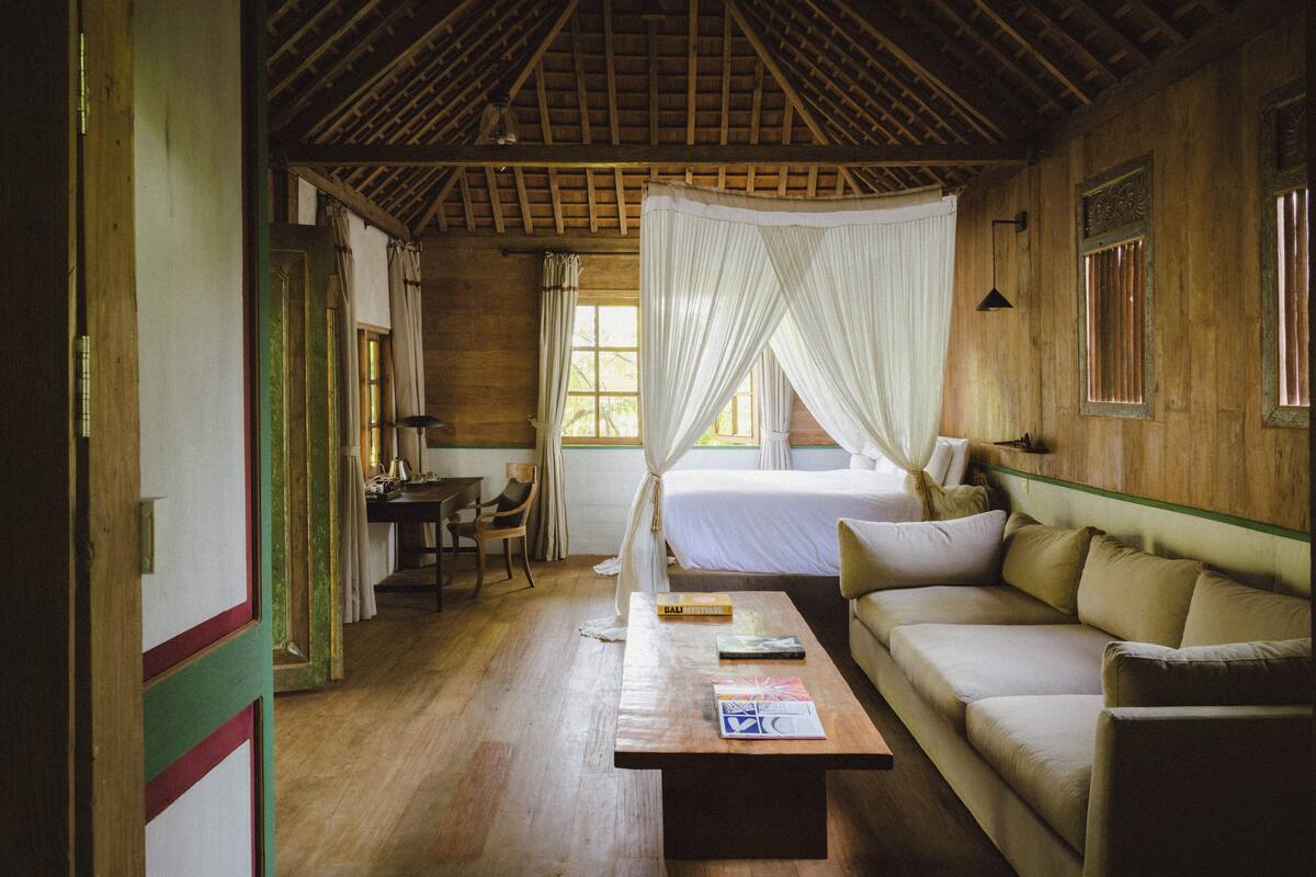 A room with a wooden ceiling, a bed with a canopy, a sofa, a coffee table with books, and a desk with a chair.