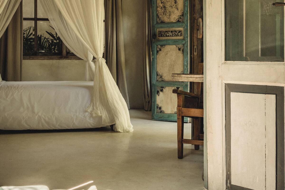 A bedroom with a bed, a wooden chair, a window with curtains, and a door with a cross.