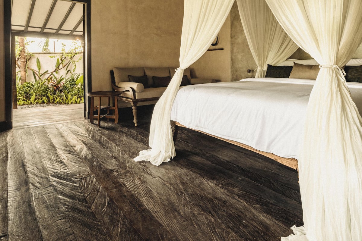 A bedroom with a four-poster bed, a wooden floor, and a view of greenery outside.
