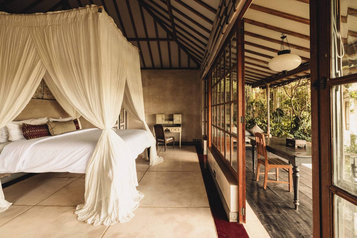 A bedroom with a canopy bed, a desk, and a view of greenery outside.