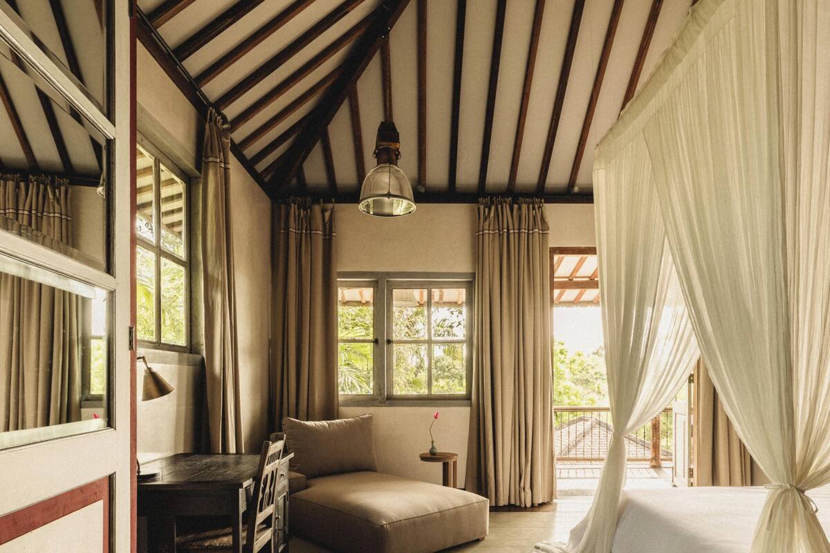 A room with a wooden floor, a bed with white curtains, a sofa, a desk with chairs, and a window with curtains.