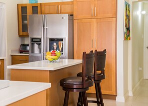 A modern kitchen with wooden cabinets, a stainless steel refrigerator, a built-in oven, and a fruit bowl on the counter. - Cozy 3BR Villa w/ Poolside Gym, AC & WiFi near Ocho Rios (Ocho Rios)
