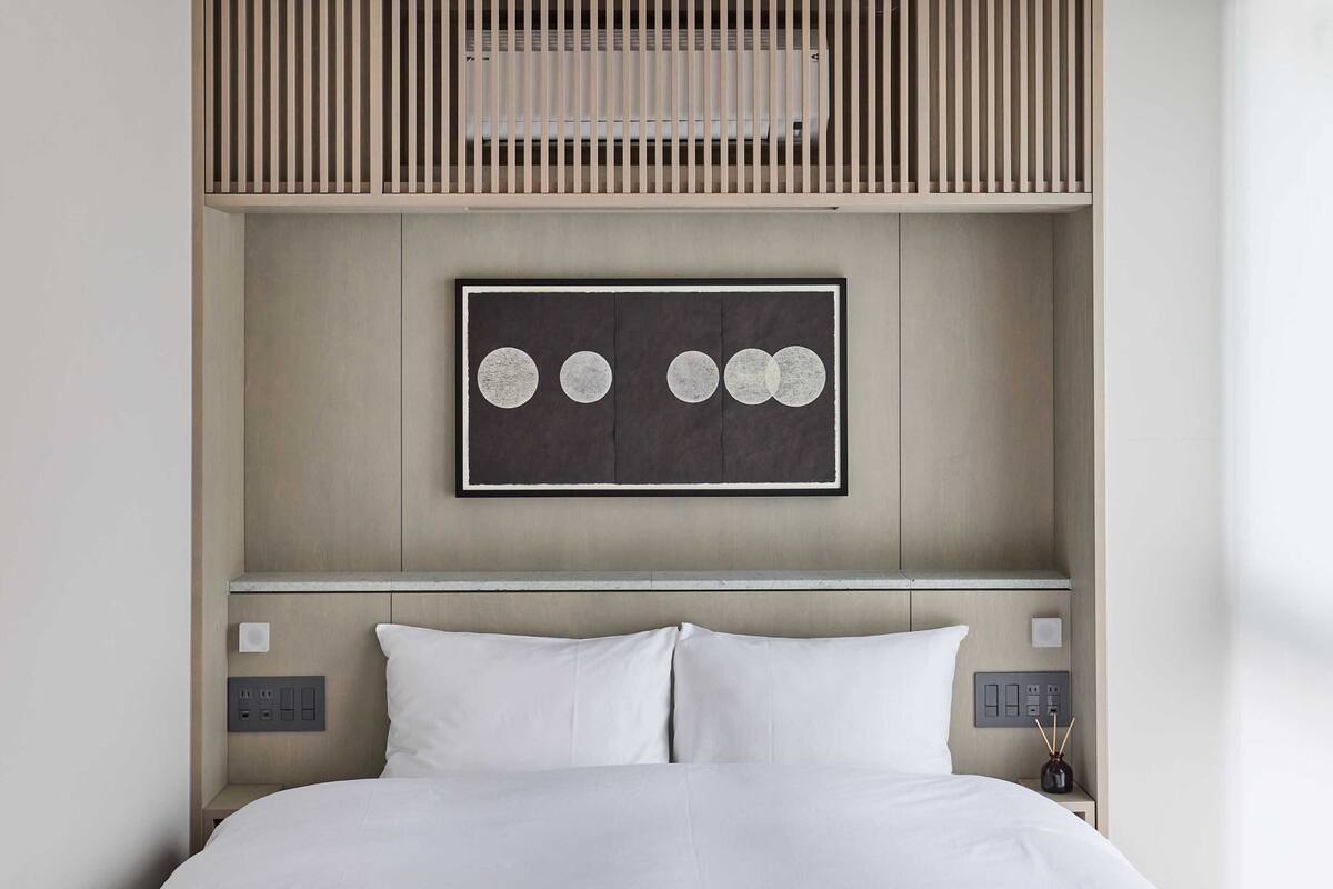 Ein modernes Hotelzimmer mit einem Bett, einem Kopfteil mit integriertem Ablagefach, einem Wandbild und Nachttischen mit Lampen.