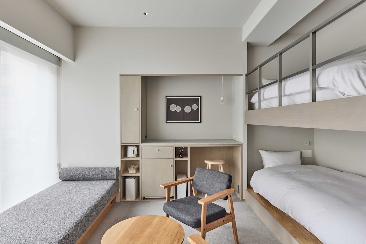 Ein kleines Zimmer mit einem Etagenbett, einem Schreibtisch mit Stuhl, einem kleinen Hocker und einem runden Tisch.