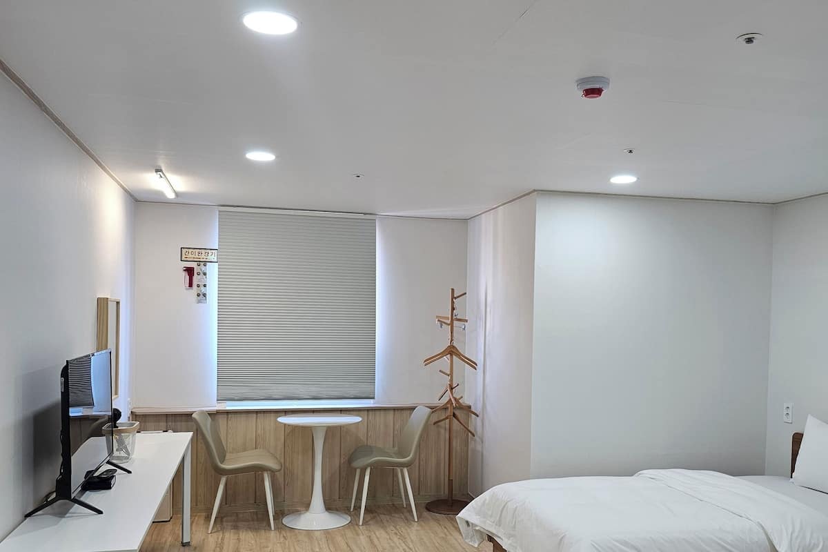 A hotel room with a desk, two chairs, a bed, and a window with blinds.