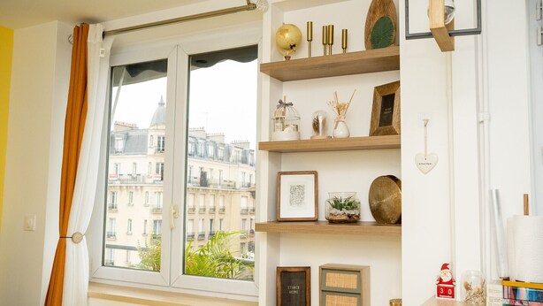 A room with a large window, wooden shelves with decorative items, a small table with books, and a view of buildings outside. - Chic Parisian Apartment - Air-Con, 1-Bed (Le Kremlin-Bicetre)