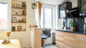 A compact kitchen with wooden cabinets, a black countertop, and a window with curtains. - Chic Parisian Apartment - Air-Con, 1-Bed (Le Kremlin-Bicetre)