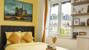 A bedroom with a bed, a painting of a cathedral, a window with curtains, and a view of buildings. - Chic Parisian Apartment - Air-Con, 1-Bed (Le Kremlin-Bicetre)