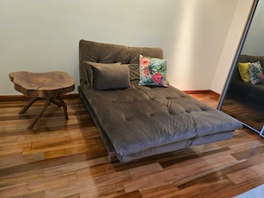 A single bed with a tufted headboard, a small wooden side table, and a mirror reflecting the room.