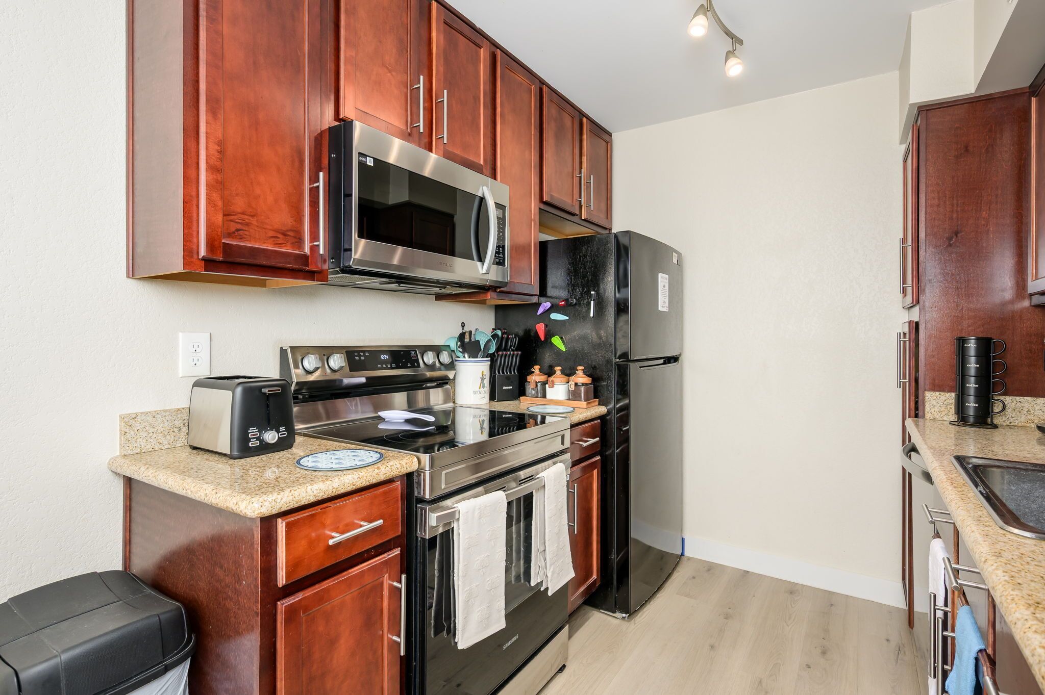 A compact kitchen with wooden cabinets, a stainless steel microwave, a black refrigerator, a toaster, and a sink.