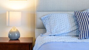 Una cama bien hecha con almohadillas estampadas, un cuadro enmarcado de un cactus en la pared y una lámpara sobre una mesita de noche de madera.