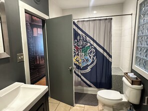 Une salle de bain avec un rideau de douche orné des armoiries de Poudlard, des toilettes et un lavabo.