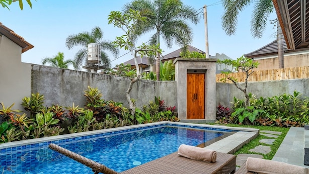 A swimming pool with lounge chairs and a wooden door, surrounded by tropical plants and a concrete wall. - Villa Deruja Ubud (Ubud)