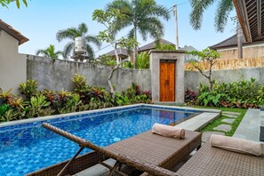 A swimming pool with lounge chairs and a wooden door, surrounded by tropical plants and a concrete wall. - Villa Deruja Ubud (Ubud)