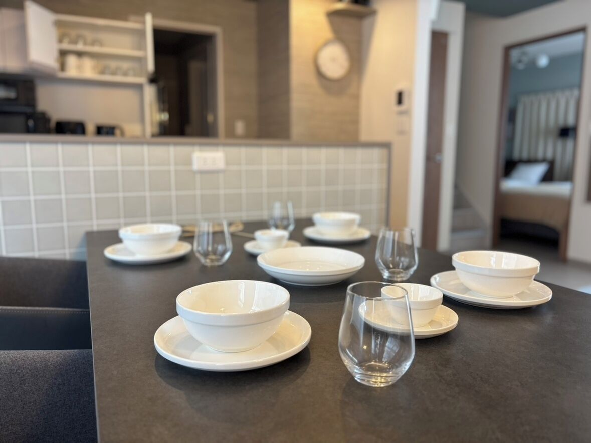 A dining table set with white bowls and plates, a glass, and a clock on the wall in the background.