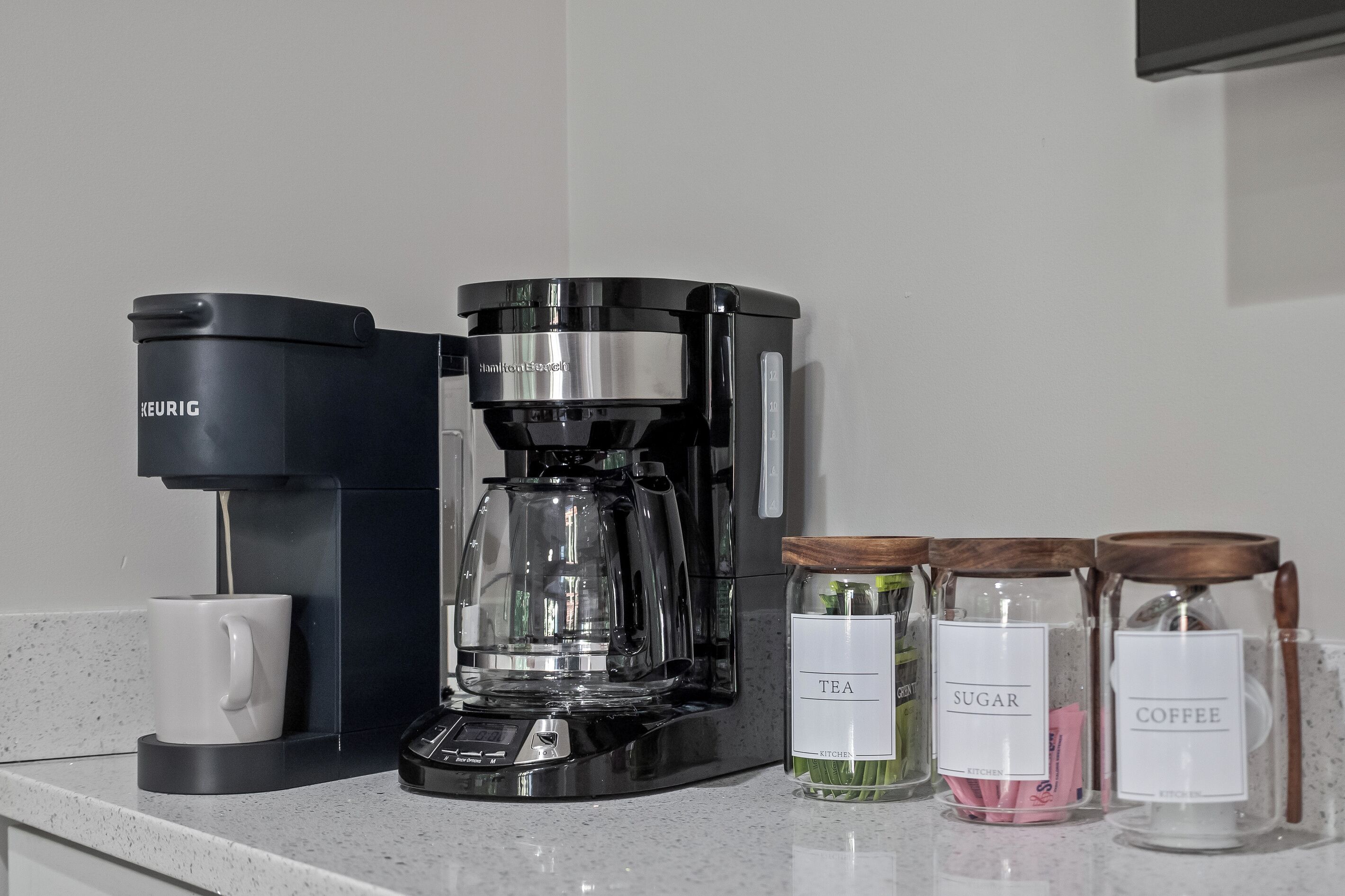 Een koffiezetapparaat, een mok en drie glazen potten met de aanduidingen thee, suiker en koffie op een aanrecht.