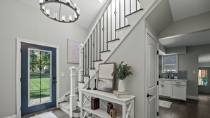 A staircase leading to a second floor, a white console table with decorative items, a door with a view of a green lawn, and a kitchen area with white cabinets.