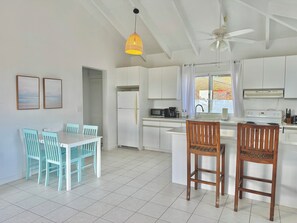 Une cuisine moderne avec des meubles blancs, un réfrigérateur blanc, un four à micro-ondes et une cuisinière. On y trouve également un coin repas avec une table blanche et des chaises turquoise.