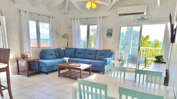 A spacious living area with a blue sofa, a wooden coffee table, and a dining table with chairs. There are large windows and a view of the outdoors.