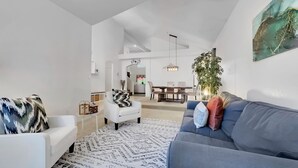 Un salon moderne avec un canapé blanc, un canapé bleu et un tapis à motifs. On aperçoit à l’arrière-plan un coin repas avec une table et des chaises.