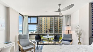 Ein Hotelzimmer mit einem großen Bett, zwei Sesseln, einem kleinen Tisch und Blick auf ein Hochhaus und einen Strand.