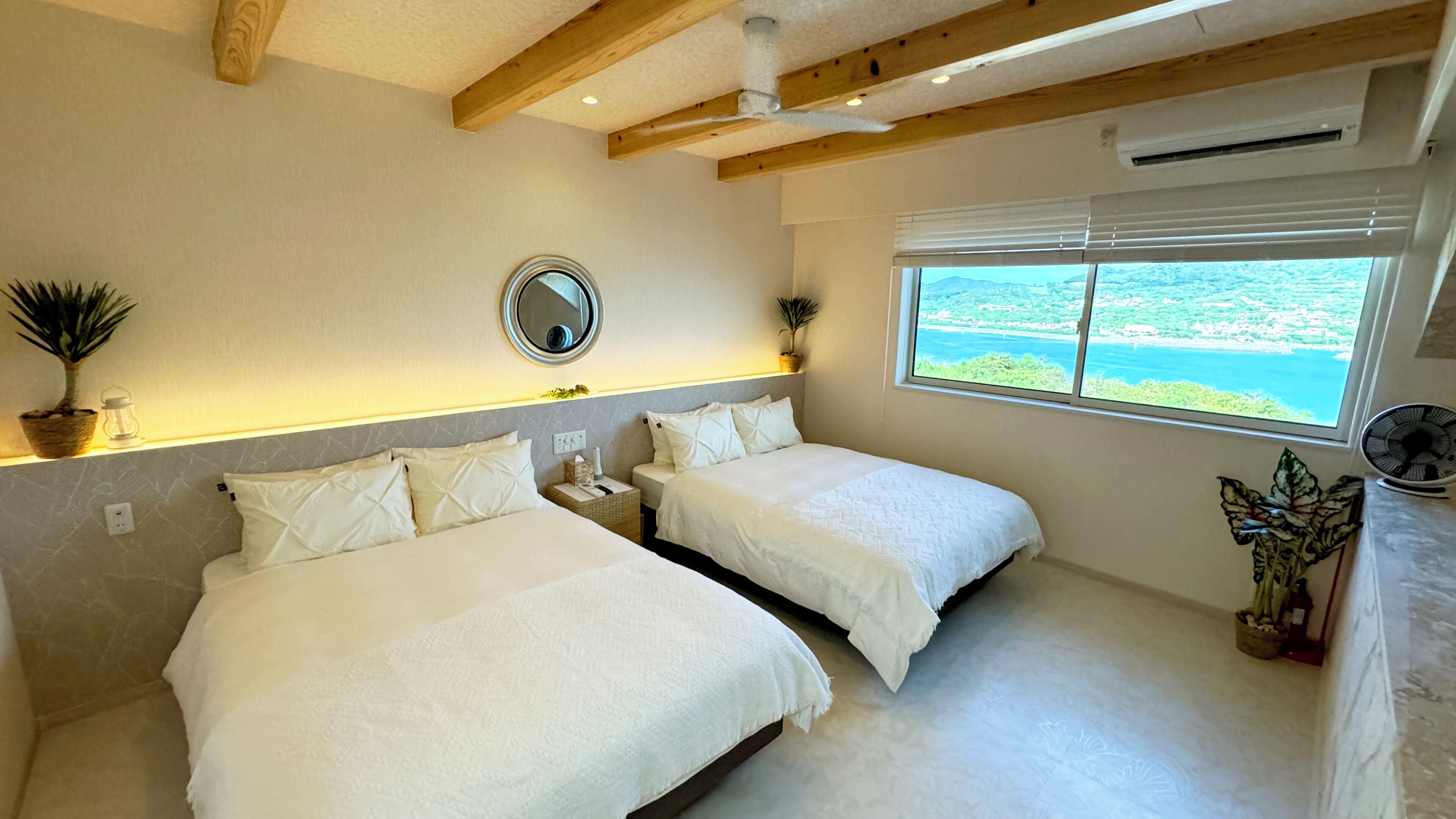 Habitación de hotel con dos camas, un ventanal con vistas, un ventilador de techo y una planta en maceta.