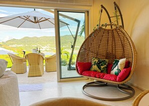 Uma cadeira suspensa de vime com almofadas vermelhas, uma mesa de vime e cadeiras, com uma vista panorâmica de colinas verdes e um corpo d'água através de uma porta de vidro.