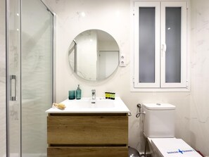 A bathroom with a white vanity, a round mirror, a toilet, and a glass shower enclosure. - Apartment + Work Area in Retiro (Madrid)