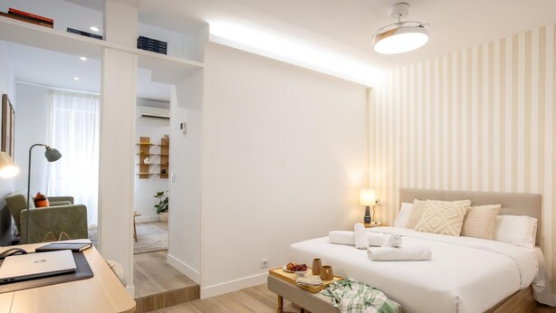 A modern hotel room with a neatly made bed, a bedside table with a lamp, a desk with a laptop, and a view of the hallway. - Apartment + Work Area in Retiro (Madrid)