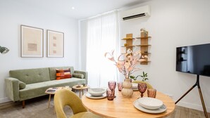 A modern living room with a green sofa, a wooden dining table with pink glassware, and a flat-screen TV mounted on the wall. - Apartment + Work Area in Retiro (Madrid)