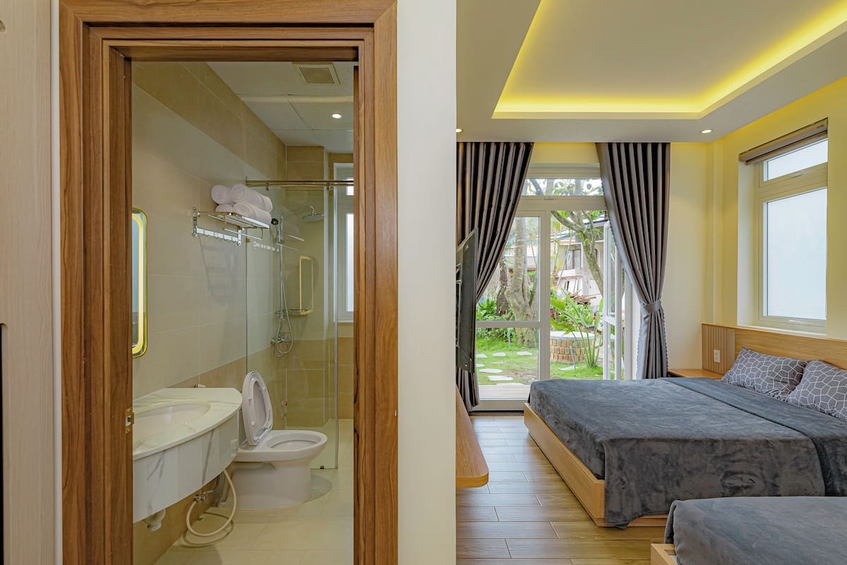 A modern bathroom with a glass shower enclosure, a toilet, and a sink. The bedroom features a large bed with a wooden headboard, a window with curtains, and a view of the outdoors.