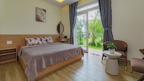 A modern bedroom with a large bed, a bedside table, a chair, and a view of greenery outside. - An Mai Boutique (Lam Vien - Da Lat)