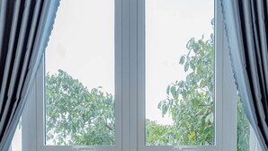 A room with two brown chairs facing a large window with a view of green foliage. - An Mai Boutique (Lam Vien - Da Lat)
