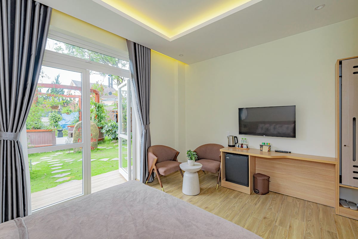 A modern hotel room with a large bed, a TV, a small table with a plant, two chairs, a desk, and a view of a garden through the sliding glass door.