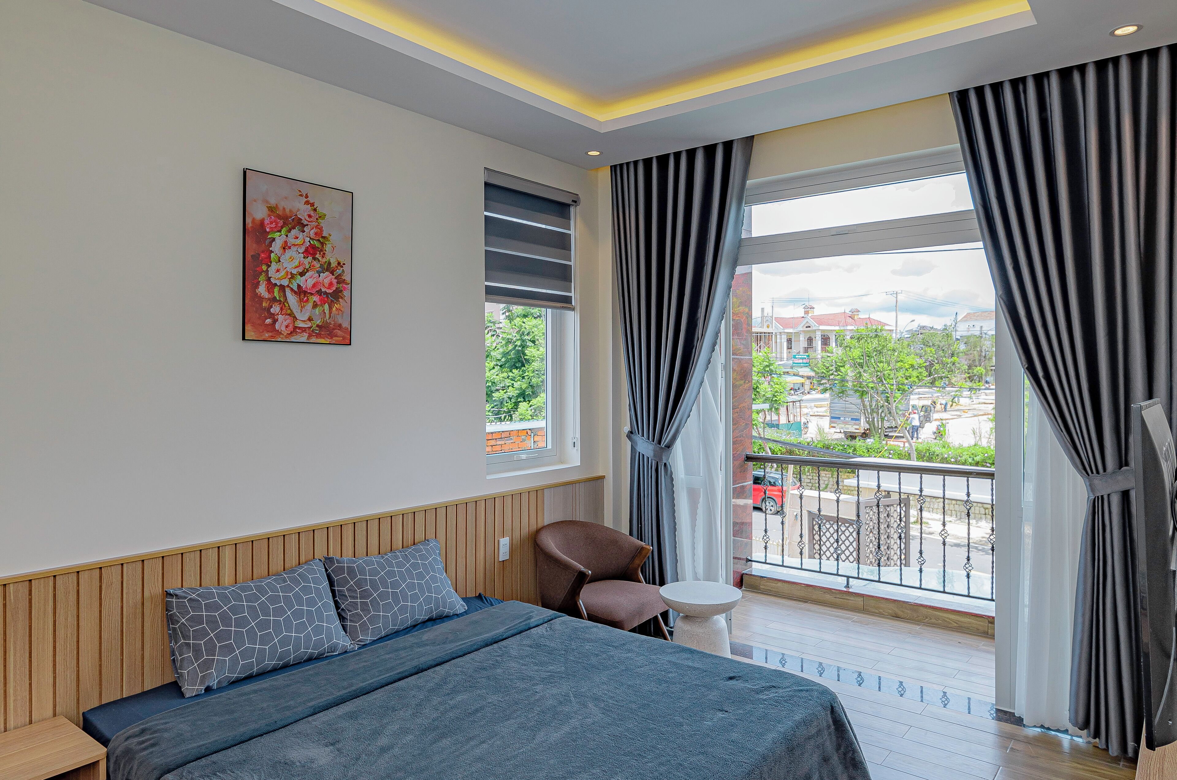 A modern hotel room with a bed, a chair, a small table, a painting on the wall, and a balcony with a view of the city.