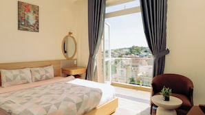 A hotel room with a large bed, a wooden nightstand, a round mirror, a small table with a plant, and a view of the city through large windows. - An Mai Boutique (Lam Vien - Da Lat)