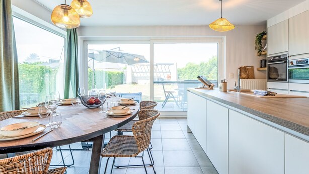 Eine moderne Küche mit weißen Schränken, einer Holz Arbeitsplatte und einem Essbereich mit Korbstühlen und einem Glastisch.
