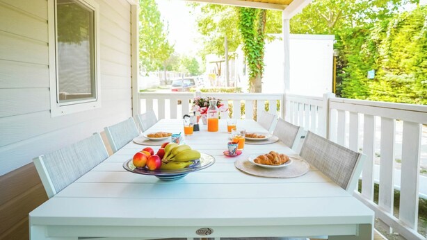 A covered outdoor dining area with a white table and chairs, a bowl of fruit, and a bottle of orange juice.