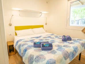 Una cama bien hecha con una colcha estampada, dos toallas enrolladas y una ventana con vista a follaje.