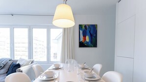 Een moderne eethoek met een witte tafel gedekt voor vier personen, een hanglamp en een kleurrijk abstract schilderij aan de muur.