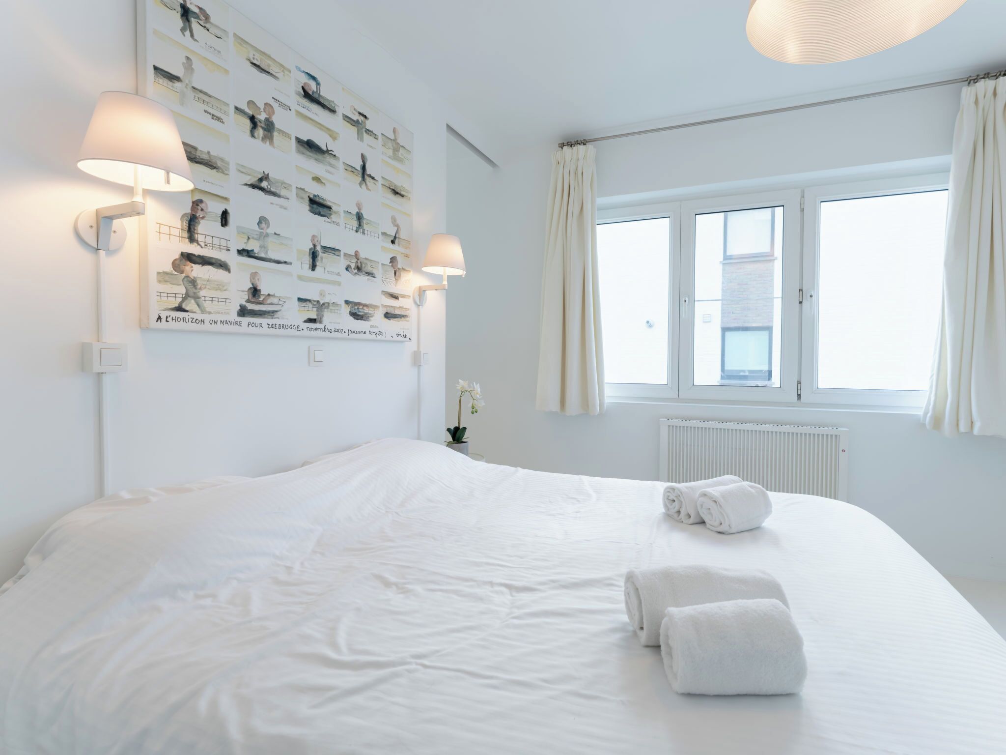 Una cama bien hecha con sábanas blancas y toallas enrolladas, una pared con múltiples cuadros enmarcados, dos lámparas de pared y una ventana con cortinas.