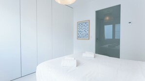 Een moderne hotelkamer met een groot bed, een hanglamp, een kunstwerk in een lijst aan de muur, en een deur die naar een andere kamer leidt.