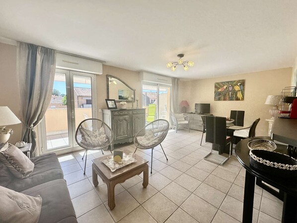 A living room with a sofa, two chairs, a small table, a TV, a painting, and a balcony with a view.