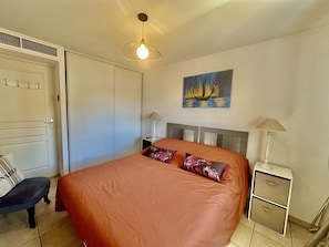 A bedroom with a bed, a nightstand, a chair, a painting, and a ceiling light fixture. - APARTMENT F2 TO RENT IN CENTER TOWN OF SAINTE-MAXIME FOR 4 PEOPLE (STE MAXIME)