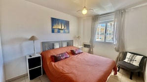 A bedroom with a bed, a nightstand, a lamp, a chair, a window with curtains, and a painting on the wall.