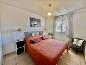A bedroom with a bed, a nightstand, a lamp, a chair, a window with curtains, and a painting on the wall.