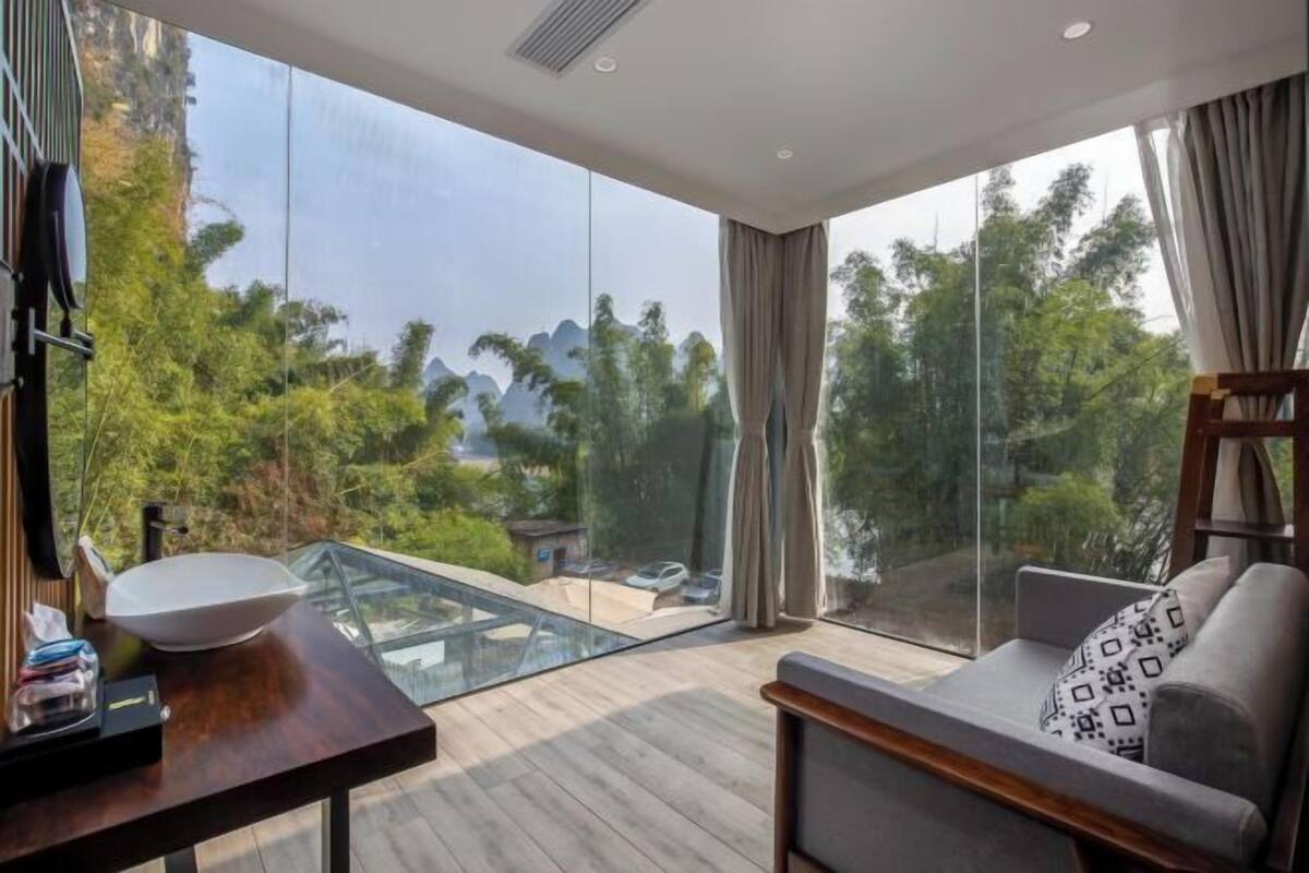 A modern room with a large glass window, a wooden table with a white bowl, a sofa with patterned cushions, and a view of greenery and mountains.